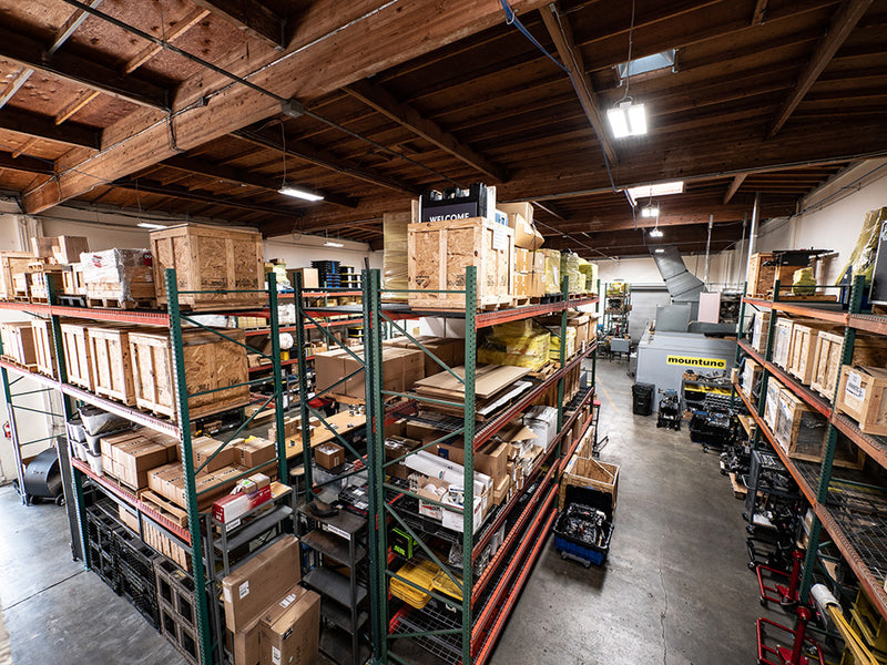Spacious warehouse interior with high shelves filled with wooden crates and various stored equipment beneath exposed wooden ceiling beams.
