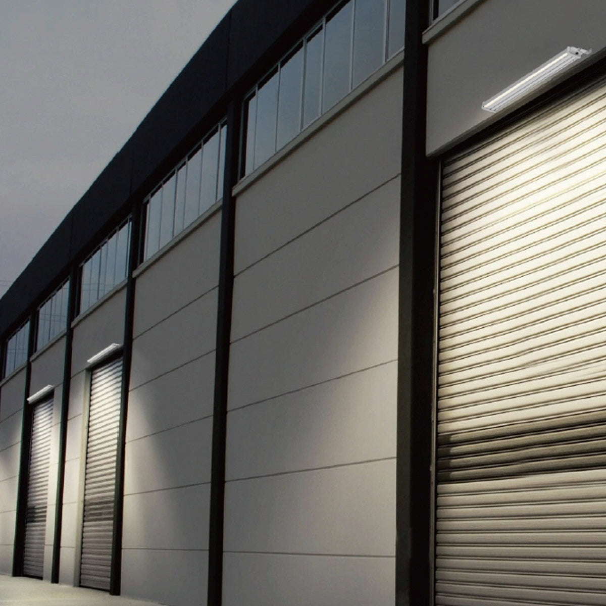 A modern industrial building facade with multiple roll-up garage doors, dark vertical framing, and linear solar lighting fixtures mounted above the doors.