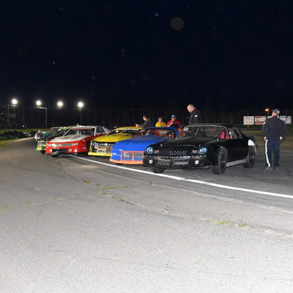 Nighttime view of a race track illuminated by LED Stadium Flood Light - 1200W with cars and crew members present.