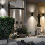 Modern outdoor wall sconces mounted on a stucco house exterior, casting warm light onto a patio with plants, a closed patio umbrella, and a chair.