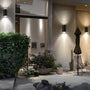 Modern outdoor wall sconces mounted on a stucco house exterior, casting warm light onto a patio with plants, a closed patio umbrella, and a chair.