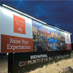 A large outdoor real estate billboard for Tesoro Viejo at dusk, featuring a white and rust-orange design with the slogan 'Raise Your Expectations' and an illustration of a resort community with pools and buildings. The bottom of the sign reads '2023 National Community of the Year. Illuminated by a run of eight solar sign lights.