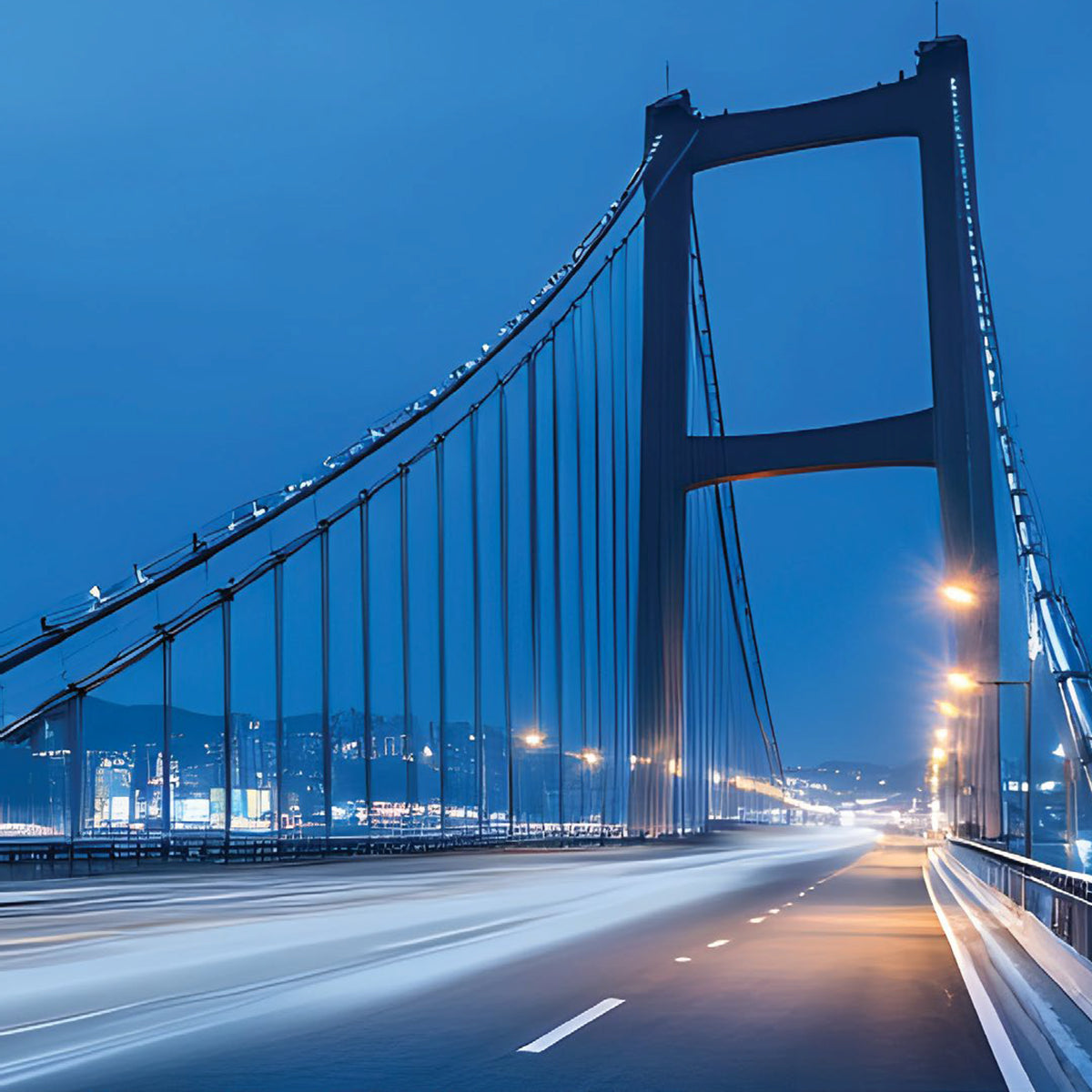 A long-exposure night photograph of a suspension bridge over a highway, with streaks of white and yellow car lights on the road and city lights visible in the distance under a deep blue twilight sky.