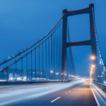 A long-exposure night photograph of a suspension bridge over a highway, with streaks of white and yellow car lights on the road and city lights visible in the distance under a deep blue twilight sky.