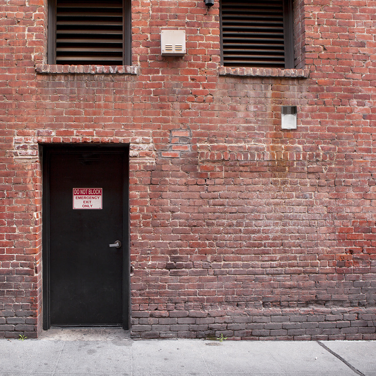 Mini LED Wall Pack w Photocell installed on brick wall above black door with emergency sign at night