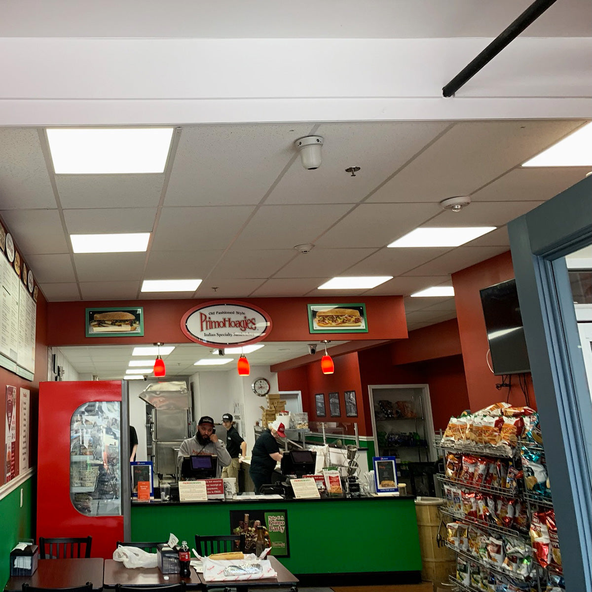 Interior of a casual sandwich shop with staff preparing orders behind the counter and snack racks on the side