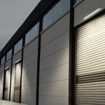 A modern industrial building facade with multiple roll-up garage doors, dark vertical framing, and linear solar lighting fixtures mounted above the doors.