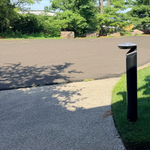 A modern black bollard light post with a curved, cap-like top mounted on a grassy verge next to a paved sidewalk, overlooking an asphalt parking lot with trees in the background.