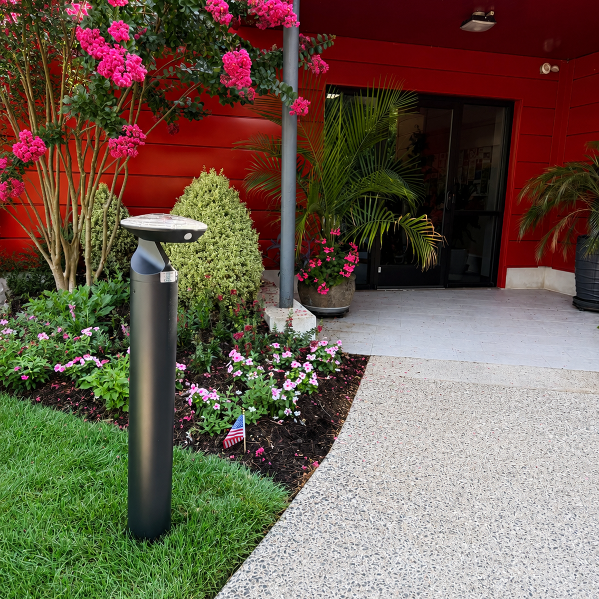 A modern black outdoor bollard light with a curved top, mounted on a grassy lawn next to a gravel path in front of a red building with pink flowers.