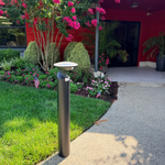 A modern outdoor bollard light post with a curved white top, installed on a landscaped lawn next to a gravel path in front of a red building.