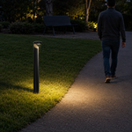 A modern black bollard outdoor light post on a grassy lawn, casting a warm yellow light onto a paved pathway. A man is seen walking along the path in the background at night.