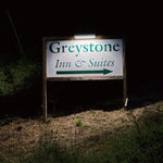 A wooden sign for Greystone Inn & Suites with green lettering and arrow, photographed outdoors at night on grassy ground, illuminated by a solar sign light placed above the sign.