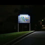 A brightly lit informational signpost at night showing a colorful map, mounted on two posts beside a grassy curb and road. Illumination provided by a top-mounted solar sign light.