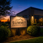 A large illuminated sign reading 'SUMMIT ENGINEERING GROUP' with a mountain logo, placed outdoors in a landscaped area in front of a modern building at dusk. Sign illuminated by a solar sign light. 
