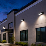 Modern exterior wall-mounted LED light fixtures installed on a light beige brick building facade with wall pack lights casting warm white light downward at dusk under a deep blue evening sky.