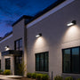 Modern exterior wall-mounted LED light fixtures installed on a light beige brick building facade with wall pack lights casting warm white light downward at dusk under a deep blue evening sky.