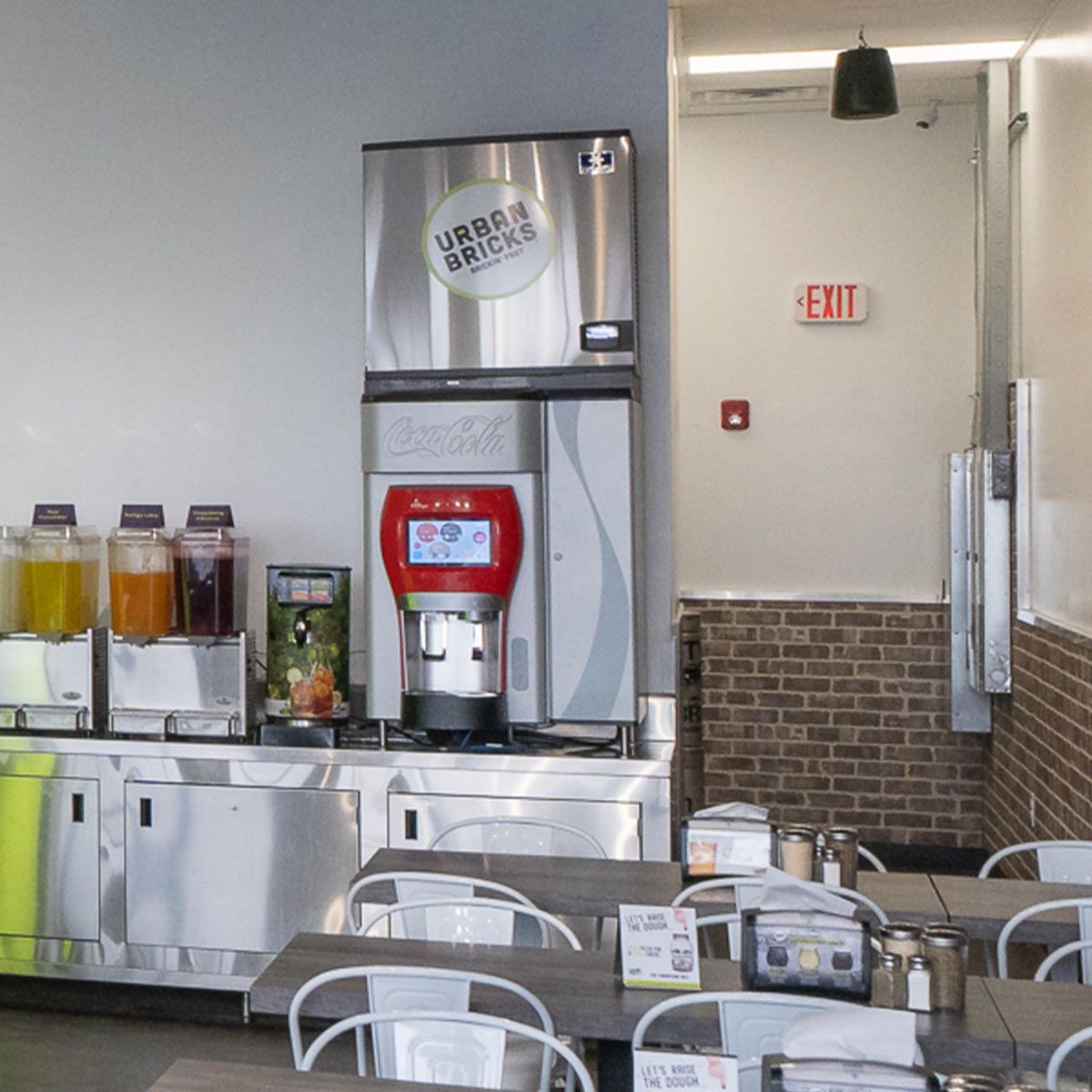 LED Exit Sign mounted on the wall of a modern cafeteria with beverage dispensers and seating area nearby