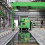Industrial facility with workers and heavy machinery under green overhead crane in a large warehouse setting