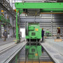 Industrial facility with workers and heavy machinery under green overhead crane in a large warehouse setting